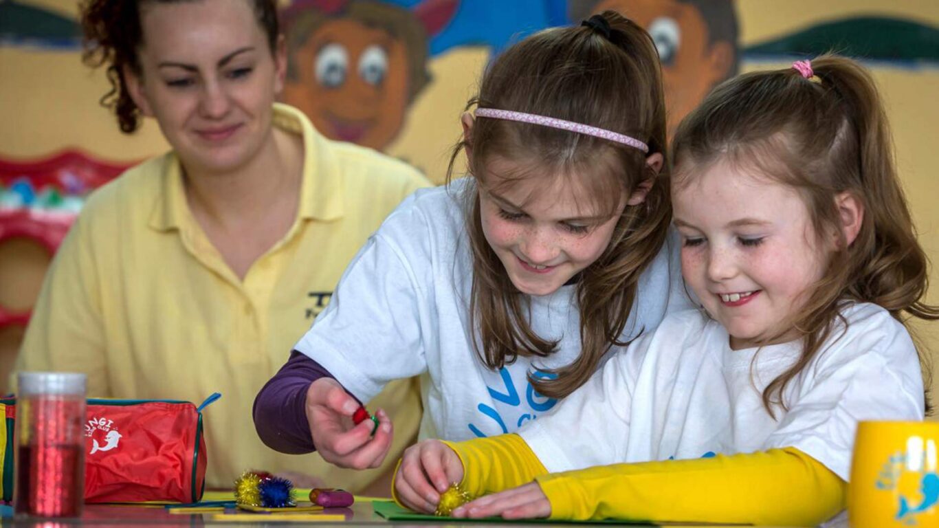 Two young girls are crafting with colorful materials at a table while an adult supervises in the background.