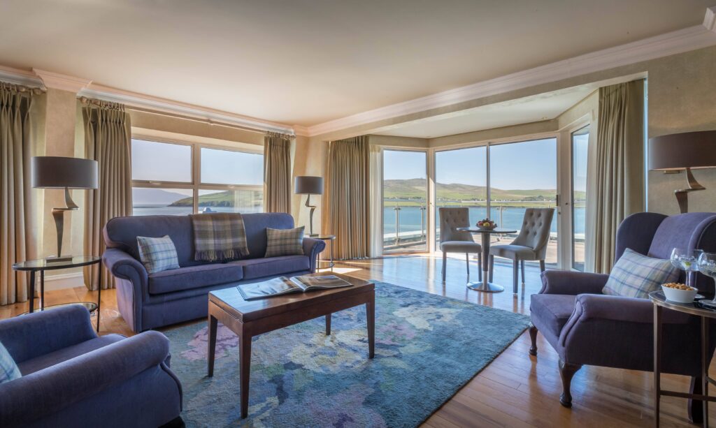 Spacious living room with large windows overlooking water and hills, furnished with blue sofas, armchairs, coffee table, and a small dining table by the window.