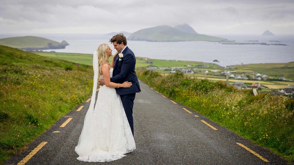 A bride and groom embrace on a rural road with a coastal landscape and green hills in the background under a cloudy sky.