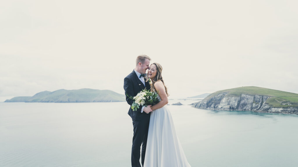A bride and groom stand together holding a bouquet, smiling, with cliffs and the ocean in the background on a cloudy day.