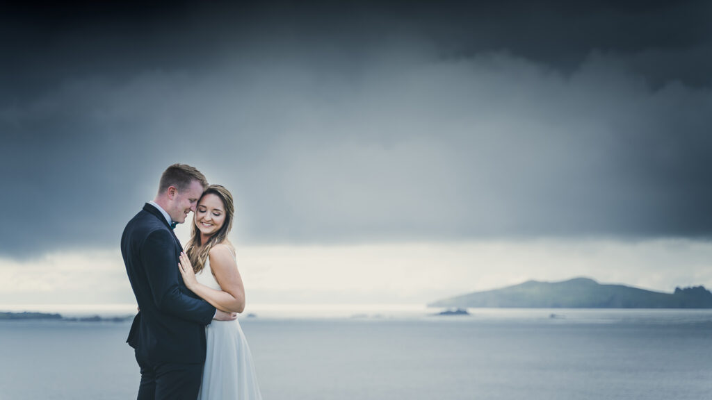 A bride and groom embrace and smile outdoors with an overcast sky and distant island visible in the background.