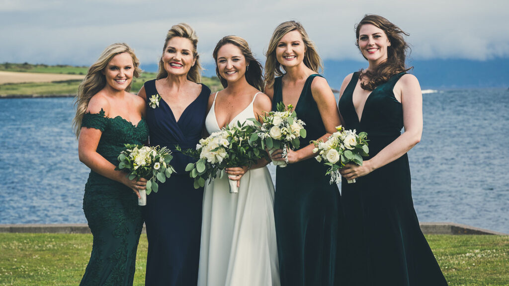 Five women in formal dresses, holding bouquets, stand together outdoors by a body of water with grassy fields and cloudy sky in the background.