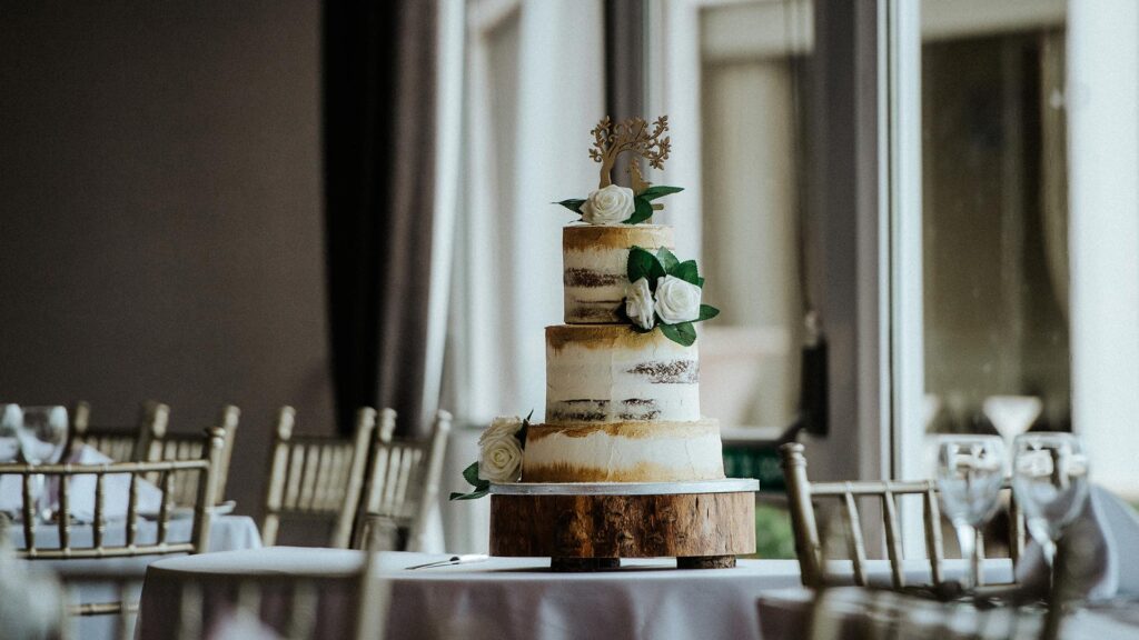 A three-tier semi-naked wedding cake with white roses and green leaves sits on a wooden stand in an elegantly set dining room.
