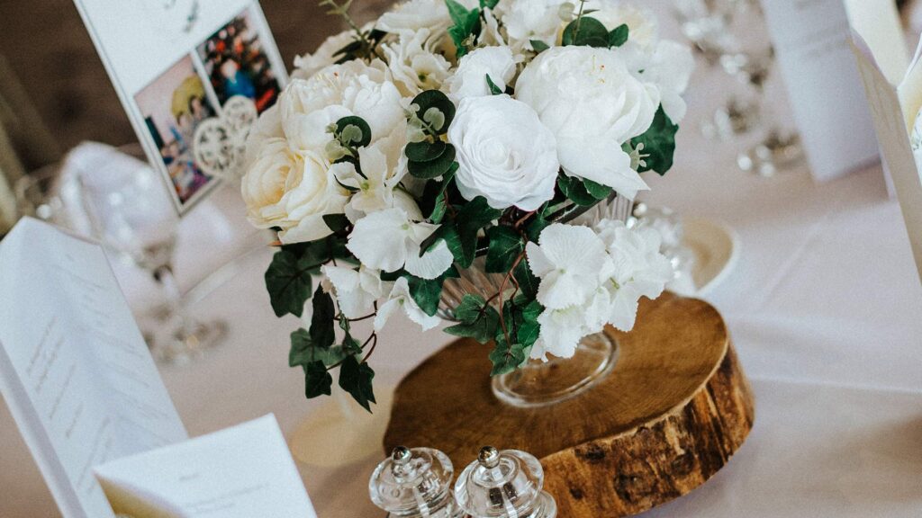 A glass vase with white roses and greenery sits on a wooden slab centerpiece, surrounded by menus and glassware on a set dining table.