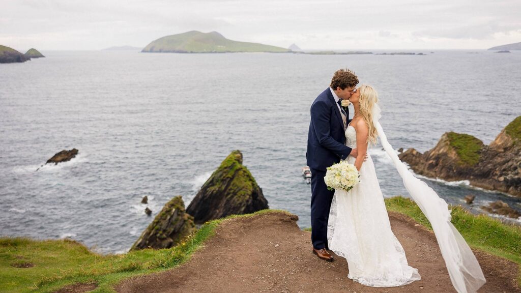 Bride and groom kiss on a grassy cliff overlooking the ocean, with rocky islands and waves visible in the background.