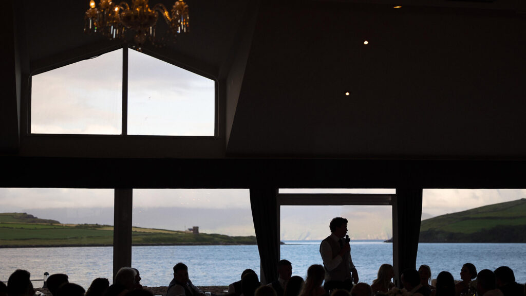 A person stands and speaks to a seated audience inside a building with large windows overlooking water and green hills.