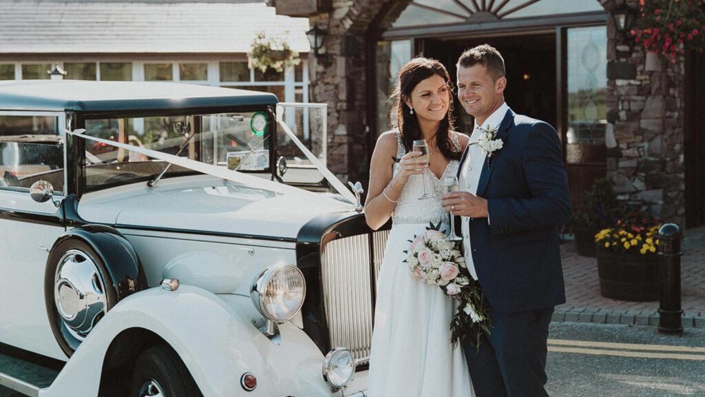 Bride and groom stand in front of a vintage white car, holding drinks and smiling, with the bride holding a bouquet of flowers.