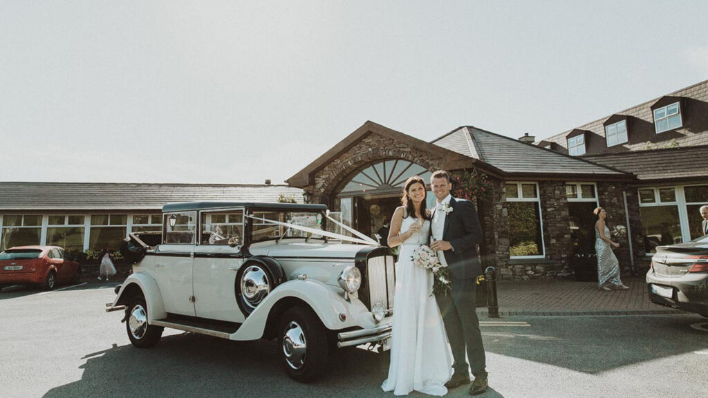 A bride and groom stand in front of a vintage white car, posing for a photo outside a stone building on a sunny day.