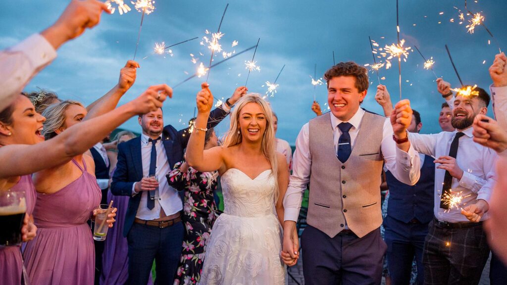 A bride and groom walk hand in hand, smiling, surrounded by guests holding sparklers at an outdoor wedding celebration.