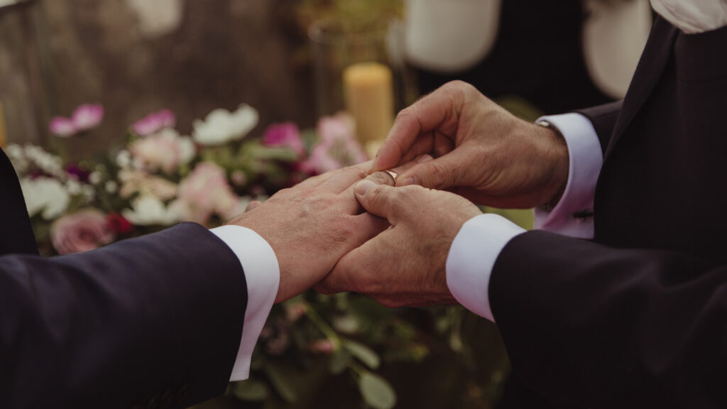A person places a ring on another person's finger during a wedding ceremony, with floral decorations visible in the background.
