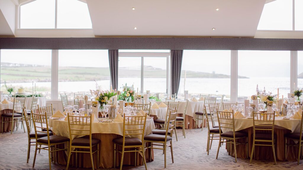 Banquet hall with round tables set for an event, featuring floral centerpieces and gold chairs, with large windows overlooking a waterfront landscape.
