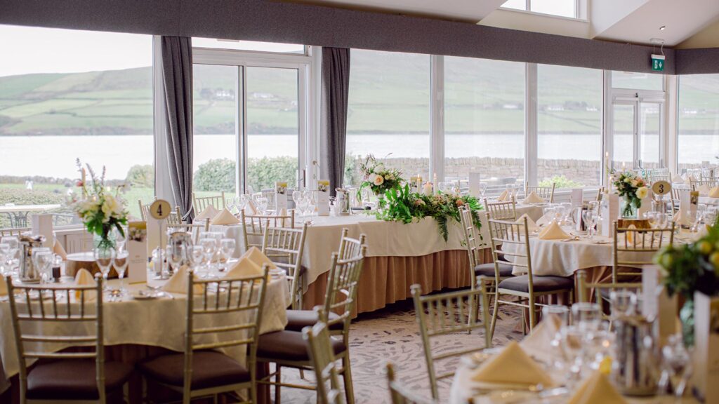 A banquet hall with round tables set for an event, featuring floral centerpieces, gold chairs, and large windows overlooking a scenic landscape with hills and water.