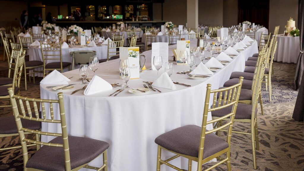 A formal banquet hall setup with long tables covered in white cloths, arranged with plates, glasses, silverware, napkins, and floral centerpieces. Gold chairs surround the tables.