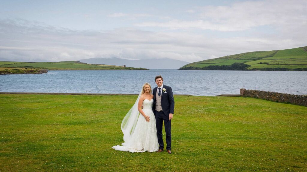 A bride and groom stand together on green grass by a lake, with hills and cloudy sky in the background.