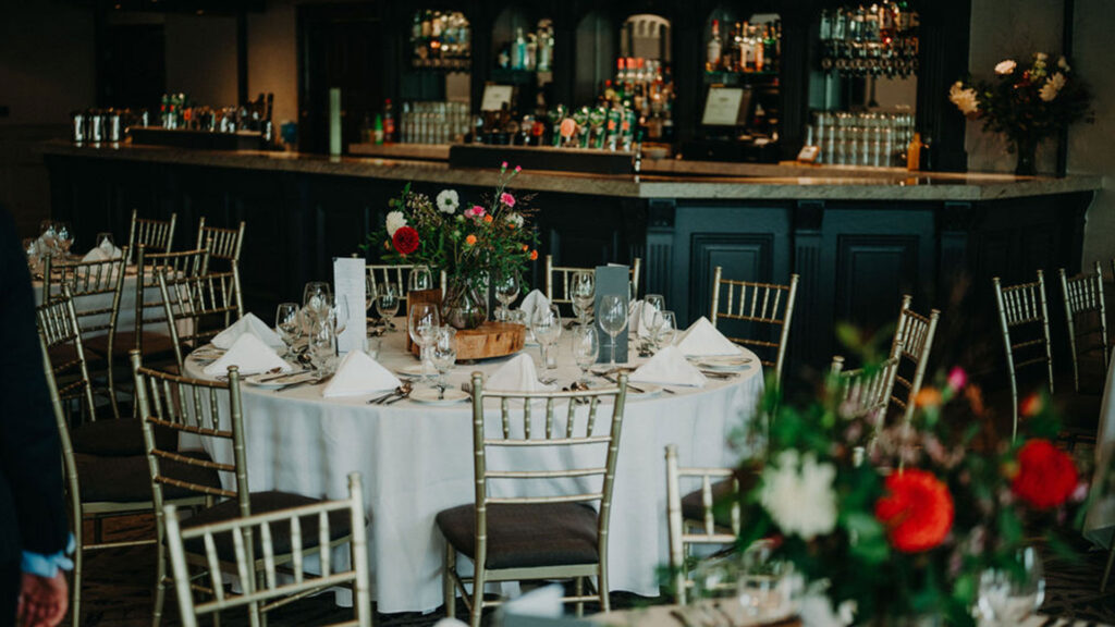 Elegant round tables with white tablecloths and floral centerpieces are set for dining in front of a fully stocked bar.