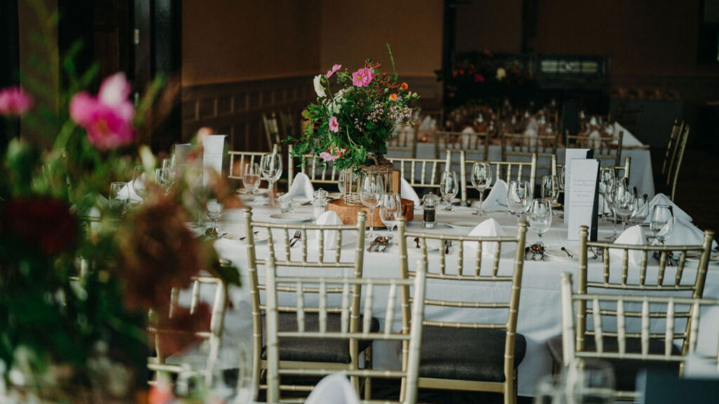 A banquet hall with tables set for a formal event, featuring white tablecloths, folded napkins, glassware, and floral centerpieces.