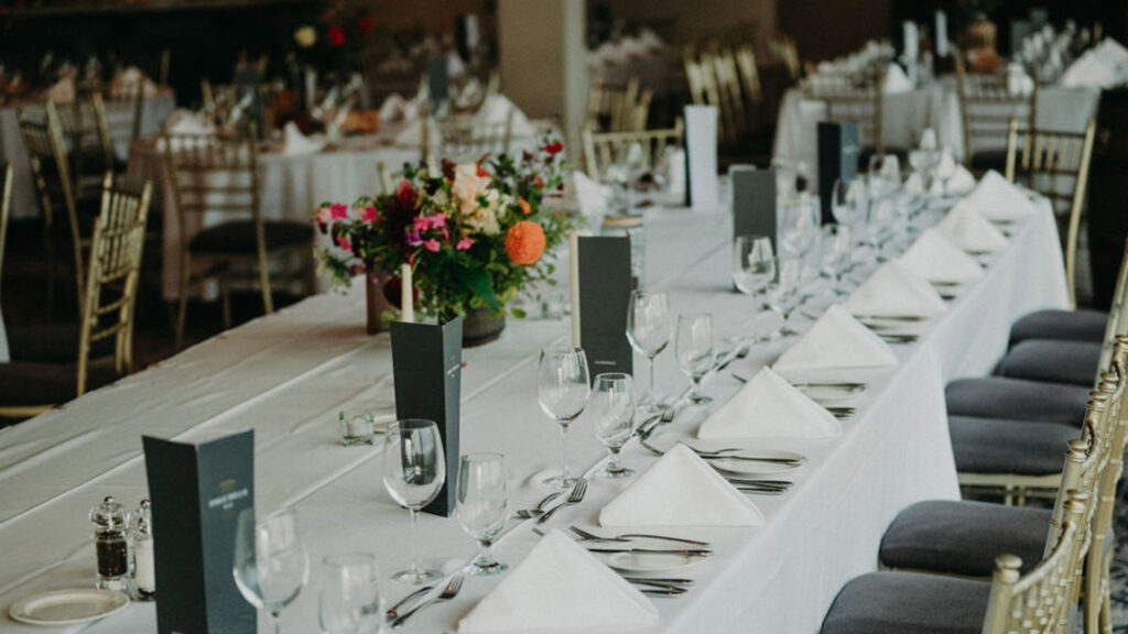 A long banquet table set with white linens, glassware, cutlery, folded napkins, menus, and a floral centerpiece in an elegant dining room.