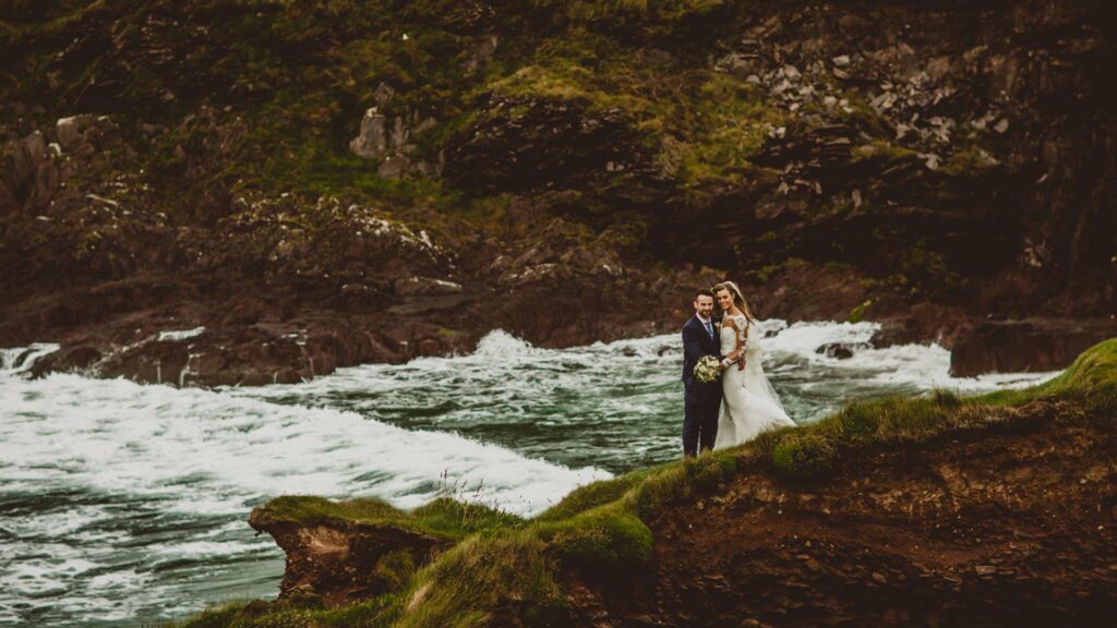 A bride and groom stand together on a grassy cliff edge, with waves crashing against rocks and a rocky hillside in the background.