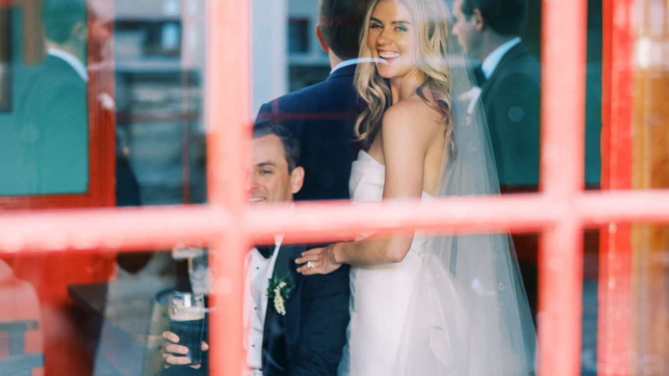 A bride in a white wedding dress stands smiling beside a seated groom, viewed through a red-paned window with other people in the background.