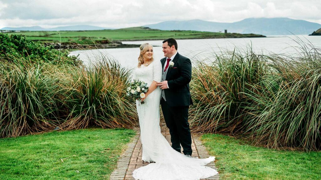 A bride and groom stand together on a path, surrounded by grass and overlooking a lake with mountains in the background.
