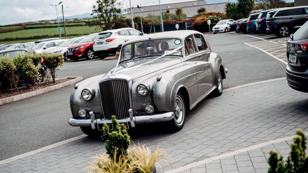 A classic silver vintage car is parked on a paved driveway, surrounded by modern vehicles in a parking lot.