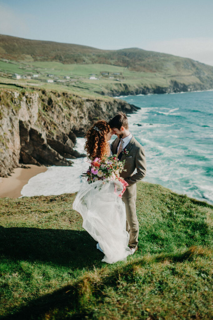 A bride and groom stand close together on a grassy cliff overlooking the ocean, with rocky shoreline and green hills in the background.