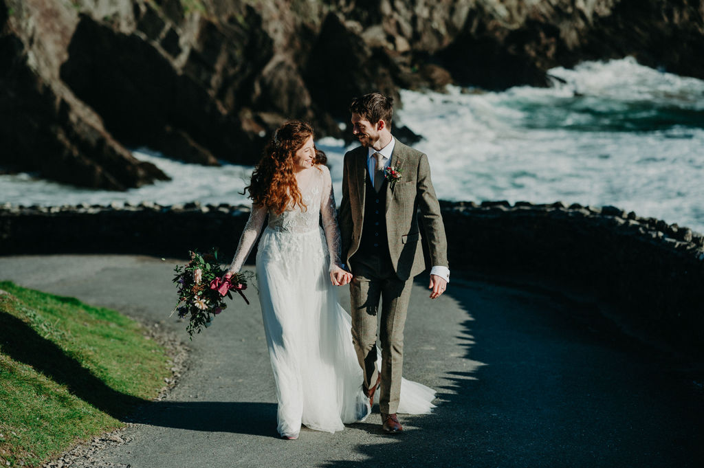 A bride and groom walk hand in hand along a seaside path, with rocky cliffs and waves in the background.