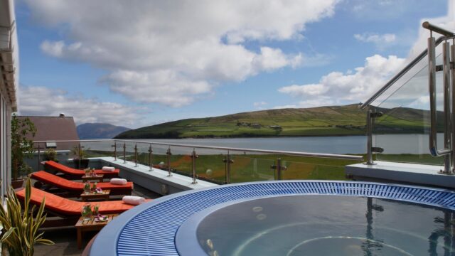 Outdoor hot tub and several lounge chairs overlook a scenic bay and green hills under a partly cloudy sky.