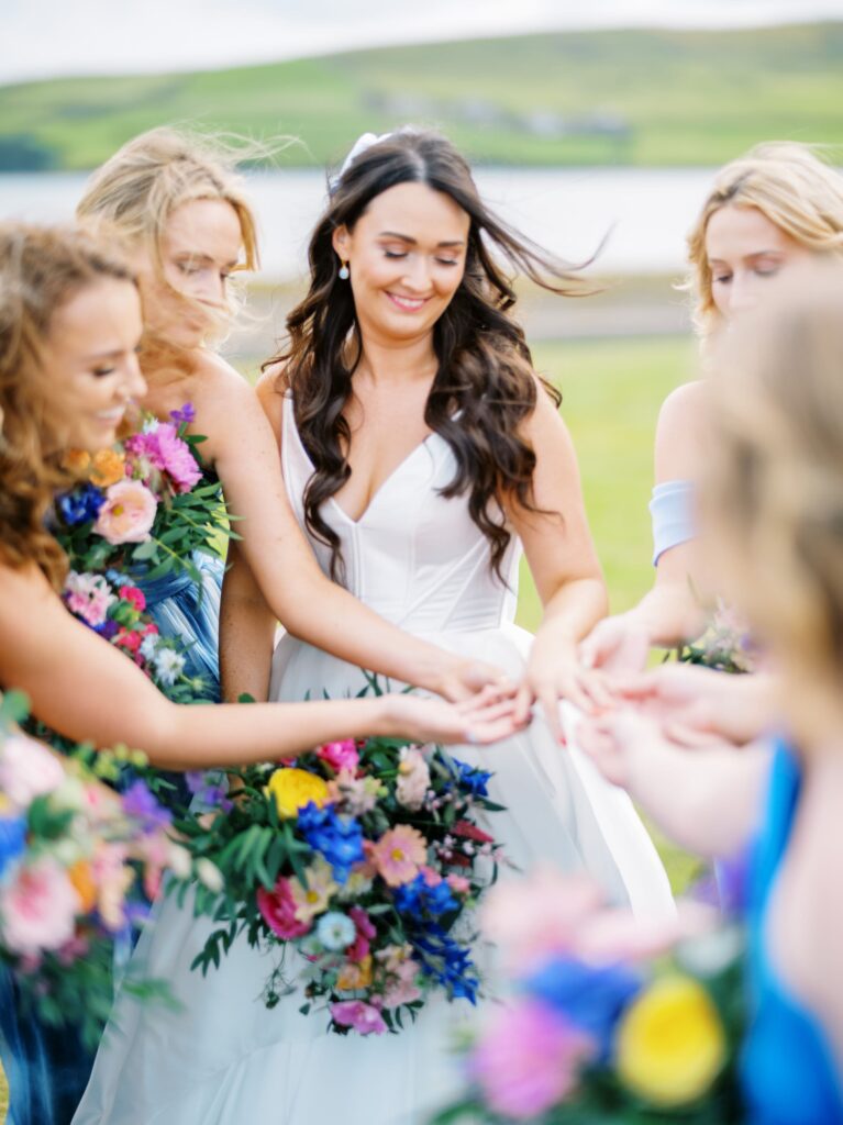 A bride in a white dress holds hands with bridesmaids holding colorful bouquets outdoors, with green hills and water in the background.