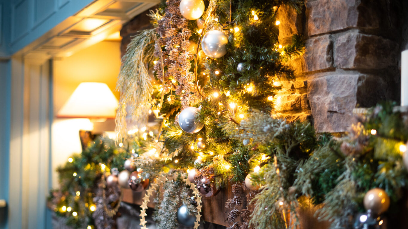 A stone fireplace mantel decorated with garlands, silver ornaments, and warm string lights, with a lit lamp in the background.