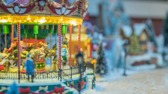 A brightly lit miniature carousel is surrounded by a snowy village scene with small houses and trees.