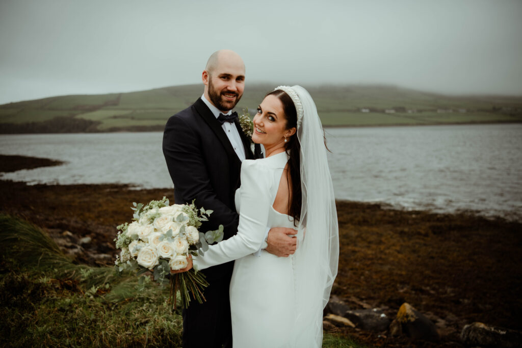 A bride and groom stand together outdoors by a lake, with the groom holding a bouquet of white flowers. The landscape in the background is green and misty.