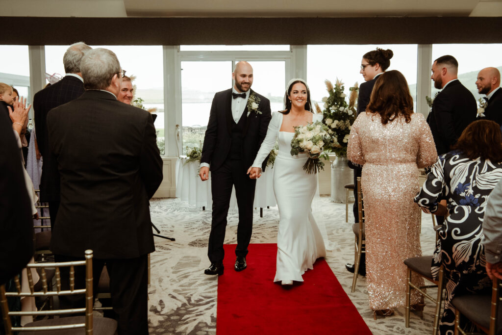 A bride and groom walk down a red aisle together, holding hands and smiling, as wedding guests stand and look on.