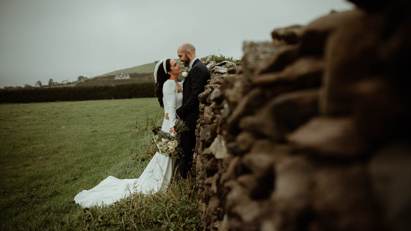 A bride and groom stand close together by a stone wall in a grassy field, with the bride holding a bouquet and wearing a long white dress.
