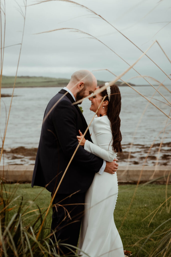 A bride and groom embrace outdoors near a body of water, standing close together and smiling on a cloudy day.