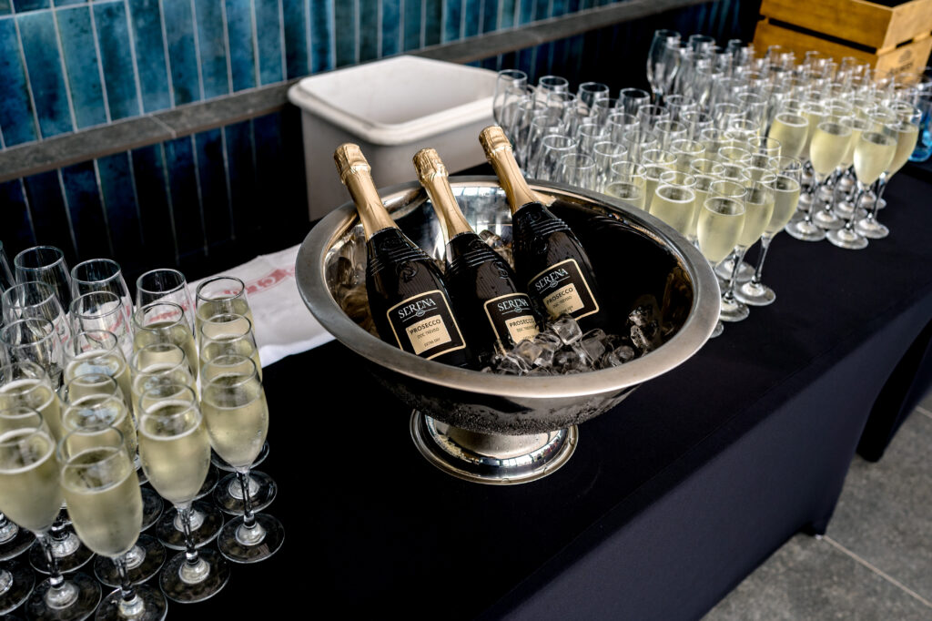 Three bottles of sparkling wine in an ice bucket are surrounded by many filled wine glasses on a black tablecloth.