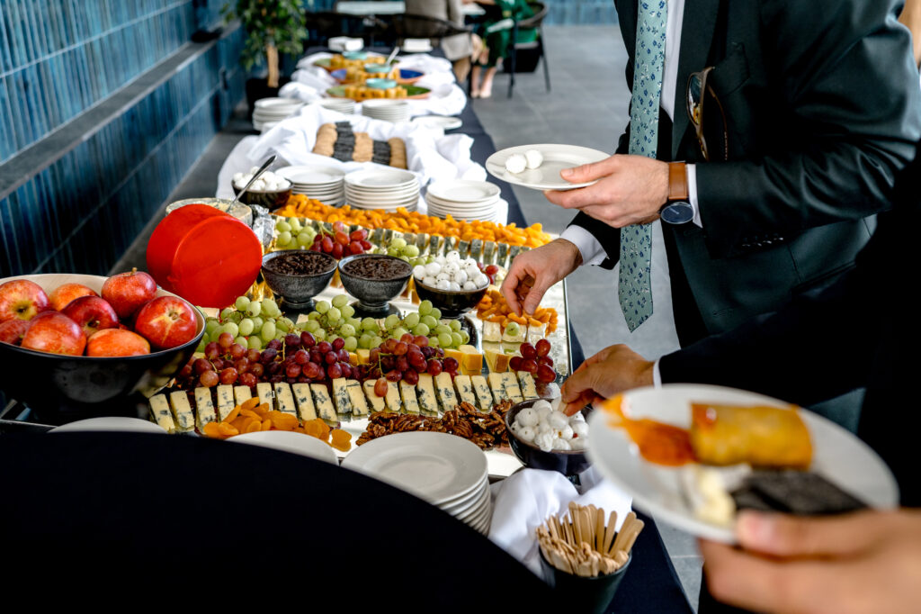 A buffet table with assorted cheeses, grapes, apples, crackers, and various snacks. People in formal attire are serving themselves food.