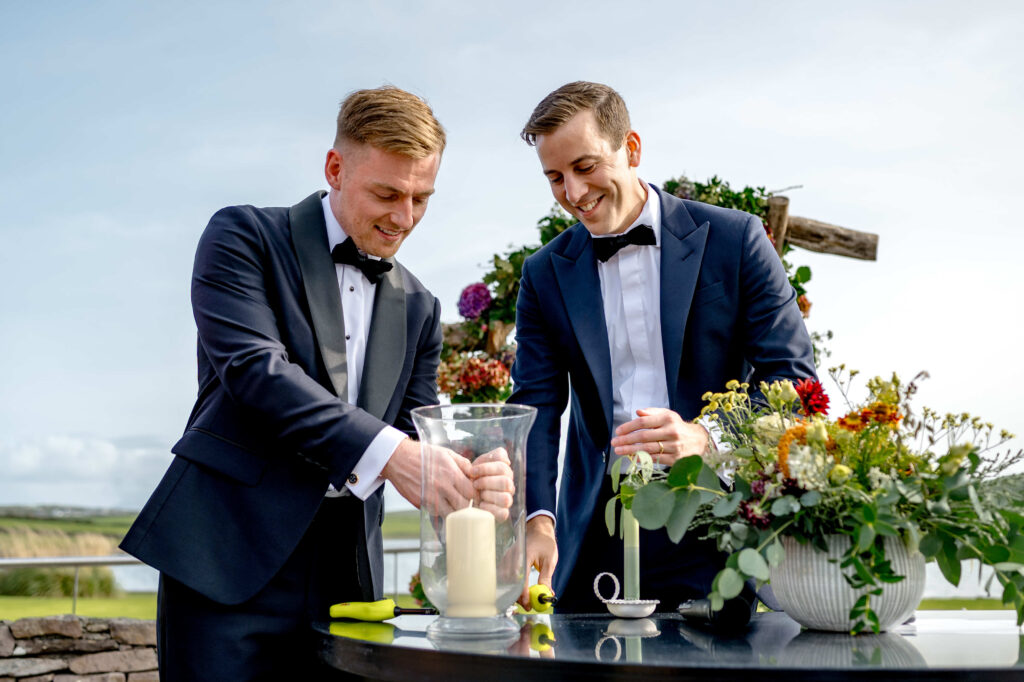 Two men in tuxedos light a large candle together outdoors, standing by a table with flowers and a green landscape in the background.