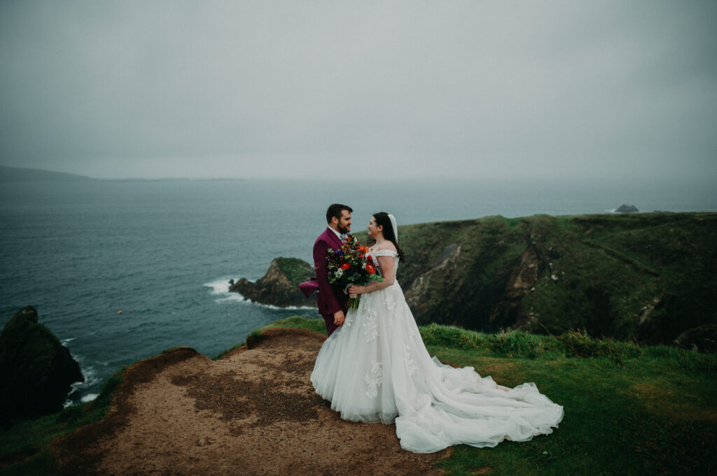 A bride and groom stand on a grassy cliffside overlooking the ocean, facing each other and holding a bouquet under a cloudy sky.