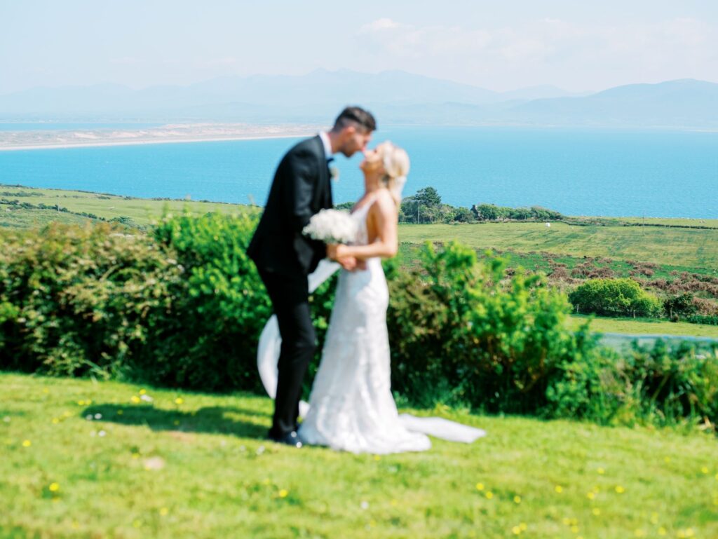 A bride and groom stand outside, kissing, with a scenic backdrop of green fields, blue sea, and distant mountains.