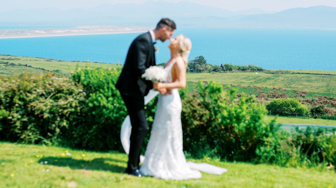 A bride and groom stand outside, kissing, with a scenic backdrop of green fields, blue sea, and distant mountains.