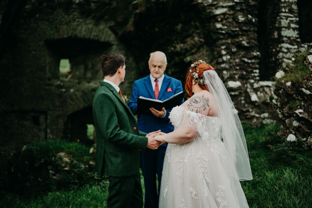 A couple holding hands during a wedding ceremony outdoors, with an officiant reading from a book in front of a stone structure.