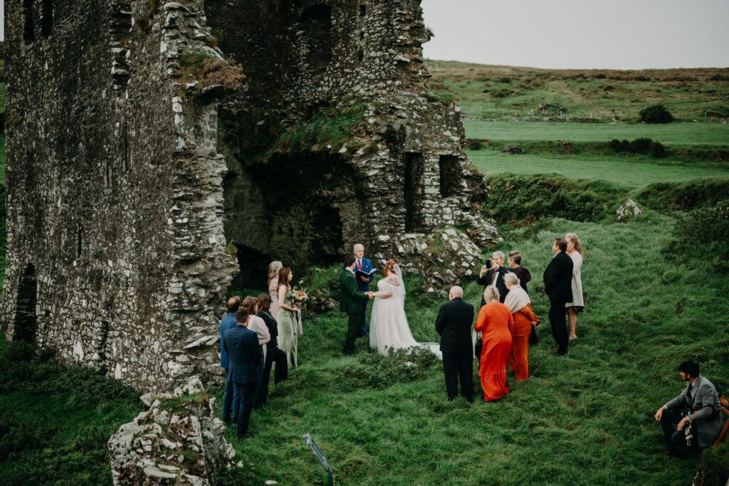 A small wedding ceremony takes place outdoors beside an old stone ruin, with the bride, groom, and guests standing on green grass.