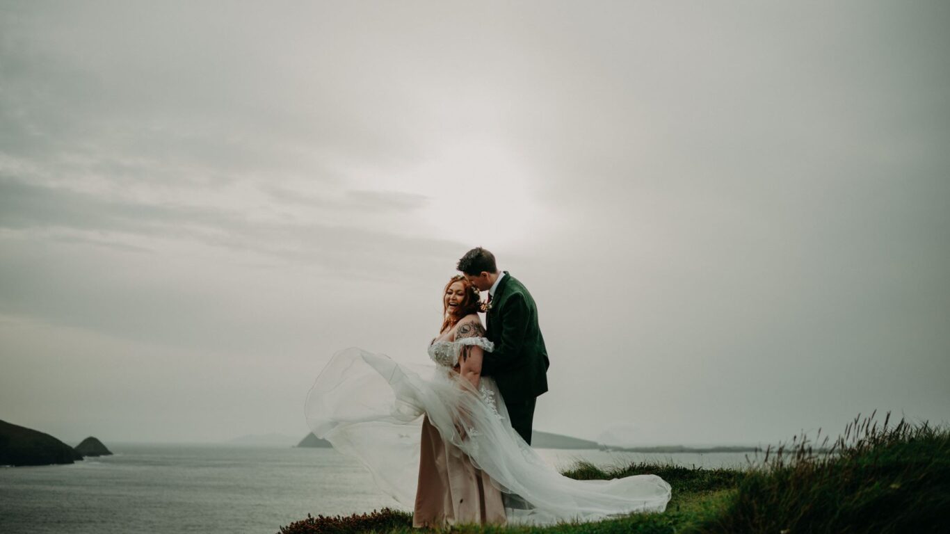 A couple in wedding attire stands on a grassy cliff with the ocean and a cloudy sky in the background. The bride's dress flows in the wind.