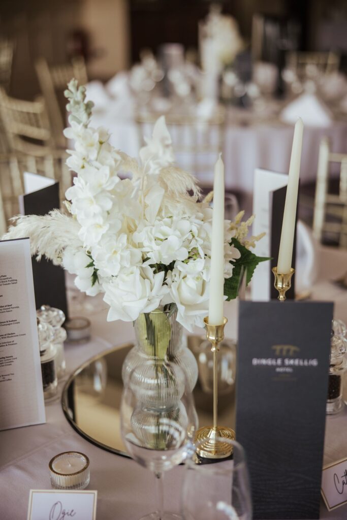 A glass vase with white flowers and two white candles on gold holders sit on a mirrored centerpiece, surrounded by menus and table place cards on a set dining table.