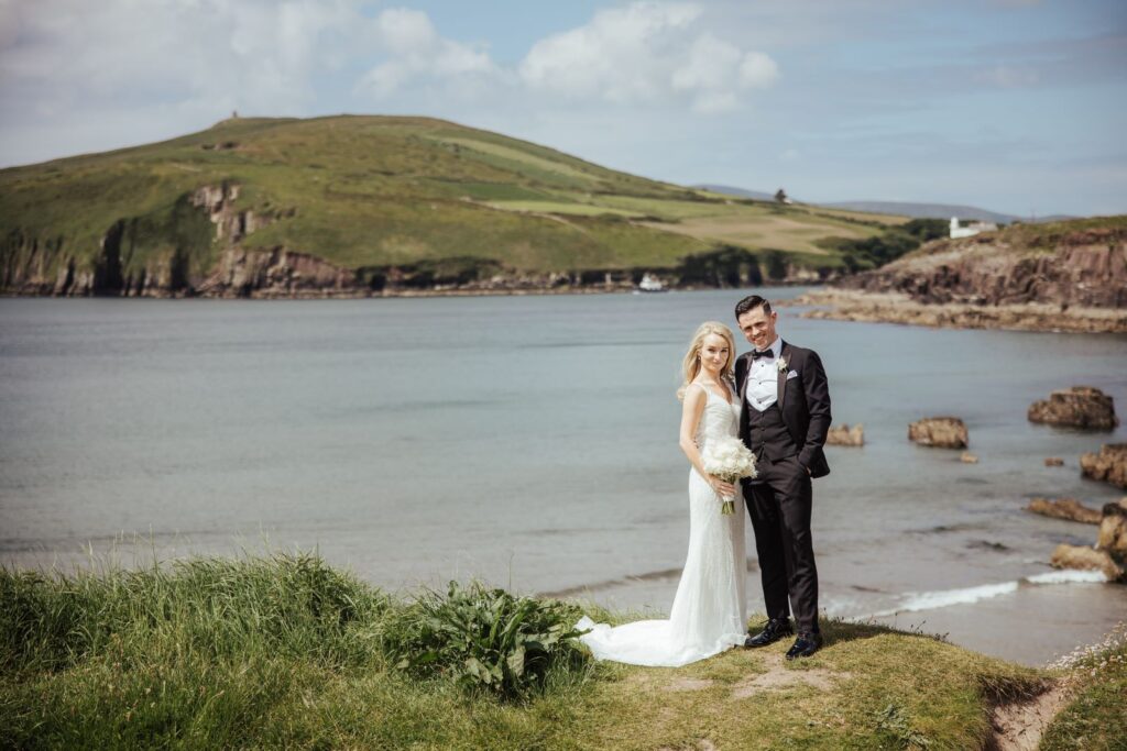 A bride and groom stand together on a grassy area by the water, with hills and cloudy sky in the background.