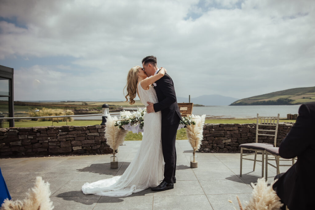 A bride and groom kiss outdoors during their wedding ceremony, standing in front of a stone wall with a scenic view of water and rolling hills in the background.