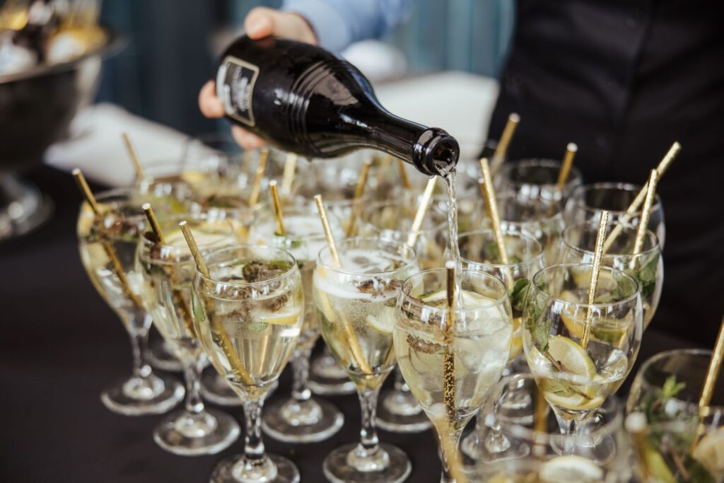 A person pours sparkling wine into multiple glasses garnished with lemon slices and herbs, each with a gold straw, arranged on a table.