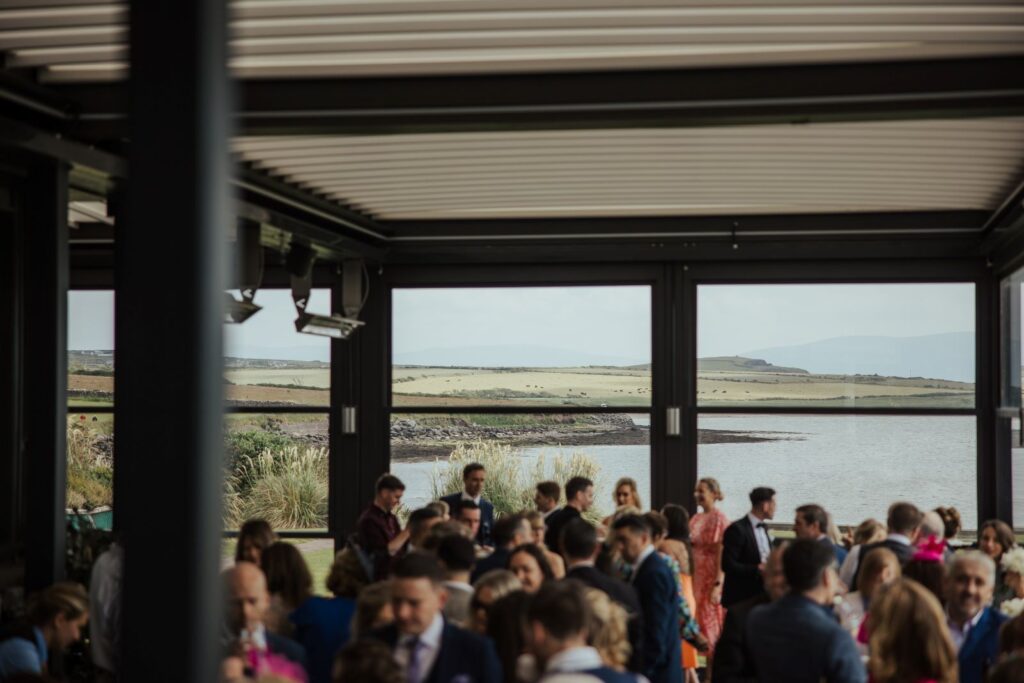 A group of people gathers inside a modern, open-sided building overlooking a scenic lake and rolling hills in the background.