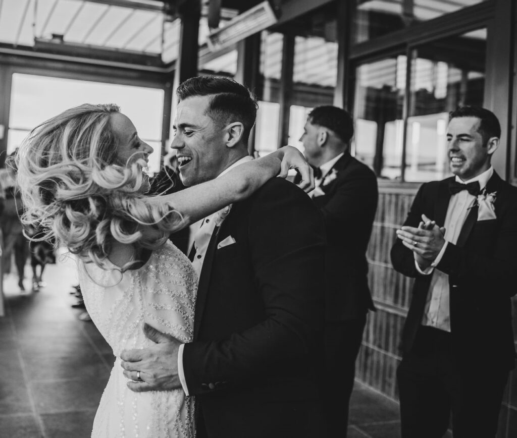A bride and groom embrace and smile at each other while two men in suits stand nearby, one clapping.